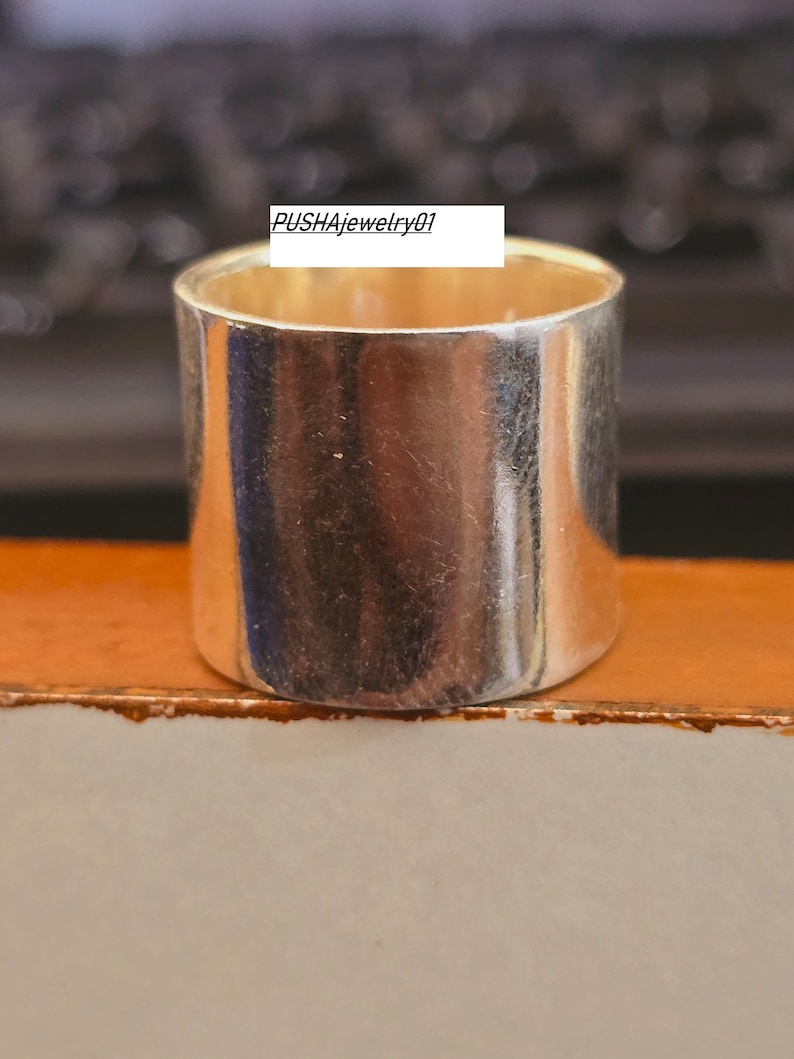 May include: A silver ring with a wide band. The ring is smooth and shiny. The text "PUSHAjewelry01" is visible on the ring.