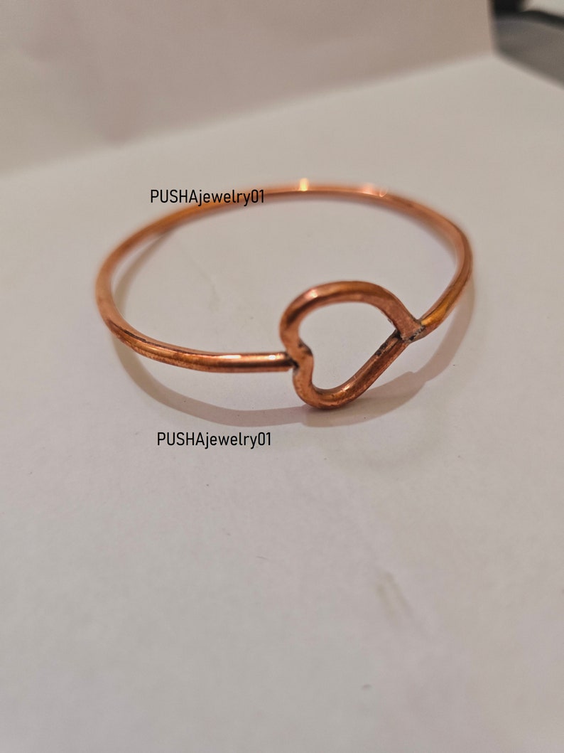 May include: A simple copper ring with a heart-shaped cutout. The ring is on a white surface.  PUSHAjewelry01 is written on the surface.
