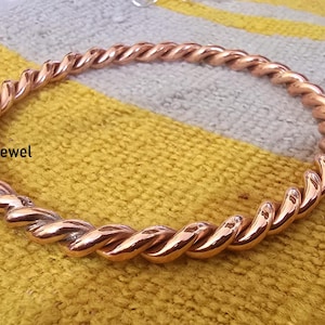 May include: A copper bracelet with a twisted design. The bracelet is on a striped fabric background.