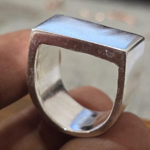 May include: A silver ring with a rectangular top and a smooth, polished finish. The ring has a modern, geometric design and a wide band. The ring is shown in a close-up shot, highlighting its reflective surface and minimalist style.