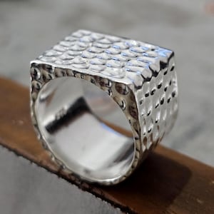 May include: A silver ring with a textured, square top. The ring features a hammered design, giving it a unique, handcrafted appearance. The band is smooth and polished, contrasting with the textured top. This piece of jewellery is suitable for everyday wear.