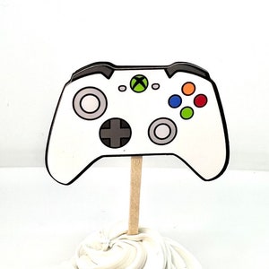 May include: A white Xbox controller cupcake topper with black trim, a wooden stick, and colorful buttons. The controller has a green Xbox logo and is placed on top of a swirl of white frosting.