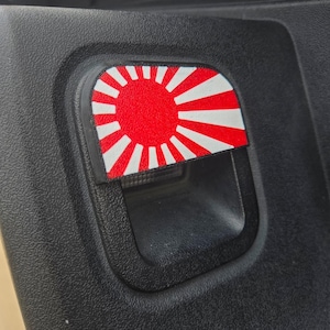 May include: A red and white Japanese rising sun flag sticker on a black car door handle.
