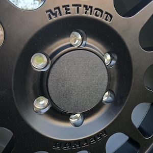 May include: Close-up of a black wheel with the word "METHOD" embossed on the rim. The wheel has a central hub cap and multiple silver lug nuts. The wheel's design includes several circular cutouts.