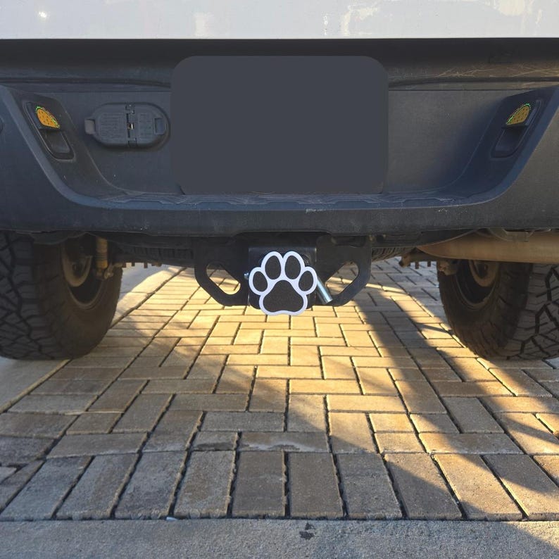 May include: A black truck hitch with a white paw print design. The hitch is attached to the back of a black truck.