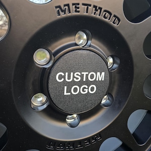 May include: Close-up of a black wheel with a silver center cap. The center cap has the text "CUSTOM LOGO" printed on it. The wheel is labeled "METHOD" on the outer rim.