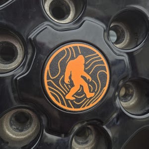 May include: A black car wheel with a round orange sticker featuring a black silhouette of Bigfoot walking on a black and orange wavy line pattern.
