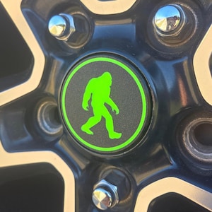 May include: A green silhouette of Bigfoot walking on a black and green wheel cover.