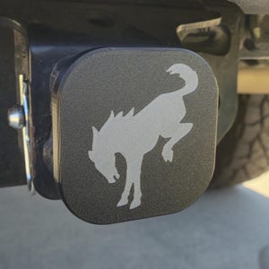 以下が含まれることがあります： 銀色の走る馬のロゴが付いた黒い金属製のヒッチカバー。カバーは正方形で、表面はテクスチャ加工されています。