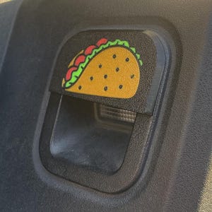 May include: A black plastic car door handle with a yellow, green, and red taco sticker on it.