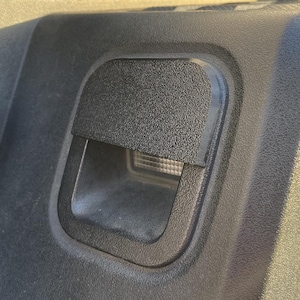 May include: A black plastic hatch with a textured surface and a rectangular opening. The hatch is likely for a light or other feature.