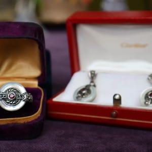 May include: A silver ring with a white center and a red stone, displayed in a purple velvet box. A pair of matching earrings in a red Cartier box. The jewelry has a vintage aesthetic.