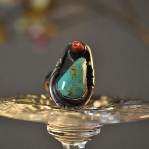 May include: A silver ring with a turquoise stone and a small red coral accent. The ring is set on a clear glass surface.