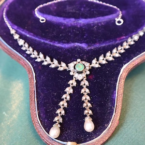 May include: A silver-toned necklace featuring a central circular green stone surrounded by small clear stones, with leaf-shaped links extending along the chain and two dangling strands ending in white teardrop-shaped pearls.