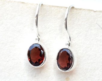 May include: A pair of silver drop earrings with oval, deep red gemstones. The gemstones are set in a silver bezel, and the earrings have a simple, elegant design. The earrings are displayed against a white background.