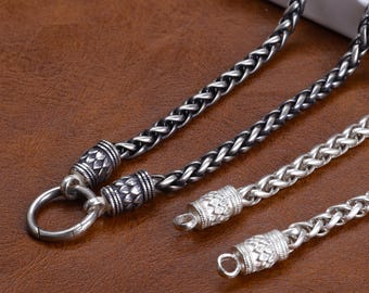 Puede incluir: Primer plano de varios collares de cadena plateados con cierres decorativos en forma de barril. Las cadenas tienen un diseño tejido y los cierres presentan patrones intrincados. Los collares se muestran sobre una superficie marrón.