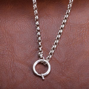 May include: A silver necklace featuring a large circular clasp. The chain is composed of small, rounded links. The clasp is open, showing a small pin. The necklace is set against a brown leather background.