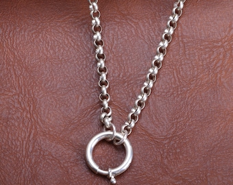 May include: A silver necklace with a large circular clasp. The chain is made of small, rounded links. The clasp is open, revealing a small pin. The necklace is displayed against a brown leather background.