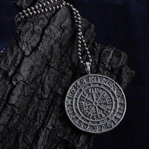 May include: A silver-toned pendant necklace featuring a circular pendant with a detailed, etched design. The pendant hangs from a ball chain. The design appears to be a compass rose or a similar navigational symbol. The background is a dark, textured surface.