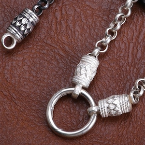 May include: Close-up of a silver chain with decorative cylindrical beads and a large circular clasp. The chain links are small and round. The beads have a detailed, textured design. The background is a brown leather-like surface.