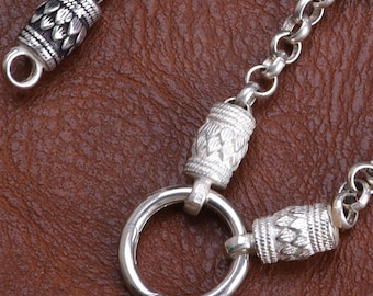 May include: Close-up of a silver chain with decorative cylindrical beads and a large circular clasp. The chain links are small and round. The beads have a detailed, textured design. The background is a brown leather-like surface.