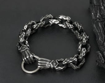May include: A silver-colored bracelet with a textured chain design. The bracelet features a clasp shaped like a hand holding a ring. The chain links have a hammered appearance, and the bracelet is set against a dark background.