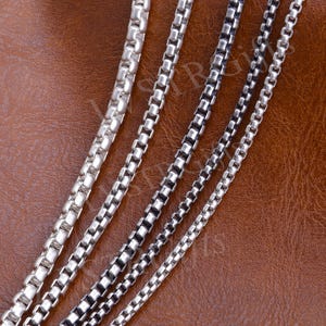 May include: Close-up of several silver box chain necklaces. The chains vary in thickness and are displayed against a brown leather-textured background. The chains are made of small, square links.