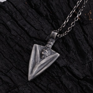 May include: A silver-toned metal pendant shaped like an arrowhead, featuring a sculpted wolf head detail at the top. The pendant is attached to a matching silver-toned metal chain link necklace with a dark, oxidized finish throughout the piece.