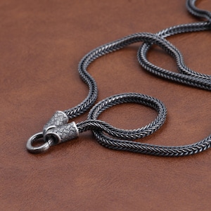 May include: Close-up of a silver-toned chain necklace with a woven design. The necklace has a circular loop and decorative end pieces. The chain is displayed on a brown surface.