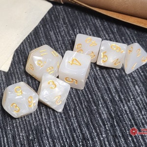 Peut inclure: Un ensemble de sept dés polyédriques d'une couleur blanc cassé semi-translucide avec une finition nacrée. Chaque dé présente des chiffres gravés remplis d'encre dorée. L'ensemble comprend diverses formes géométriques pour les jeux de rôle.