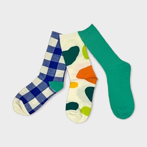 Könnte beinhalten: Drei Paar Socken, eines mit einem blau-weiß karierten Muster, eines mit weißem Hintergrund und bunten abstrakten Formen und eines einfarbig grün.