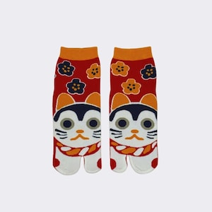 May include: A pair of red and white tabi socks with a cartoon cat design. The cat has orange ears, black eyes, and a white face. The socks have a floral pattern with orange, black, and white flowers.