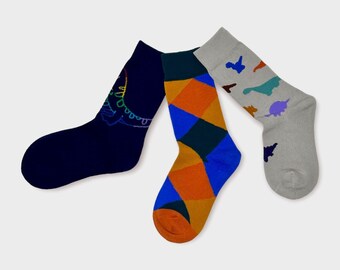 Puede incluir: Tres pares de calcetines coloridos. Un par es azul marino con un diseño de arcoíris, otro es azul y naranja con un patrón geométrico, y el último par es gris con un estampado de dinosaurio.