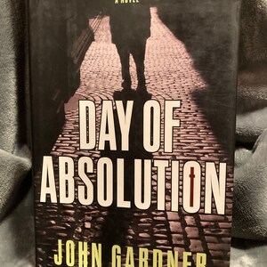 Puede incluir: Una portada de libro en blanco y negro con el título "Day of Absolution" en letras blancas grandes. El nombre del autor, "John Gardner", está impreso en letras amarillas debajo del título. La portada presenta la silueta de una persona caminando por una calle adoquinada.
