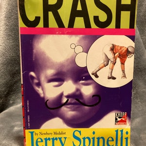 May include: A paperback book cover with a yellow background and the title "CRASH" in black letters. The cover features a cartoon illustration of a baby with a mustache and a football player in the background. The author's name, "Jerry Spinelli", is written in blue letters below the title. The book is by Newbery Medalist Jerry Spinelli.
