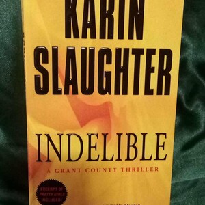 May include: A yellow and orange book cover with the title "Indelible" in black text. The author's name, "Karin Slaughter", is in large black text above the title. The book is a Grant County thriller.