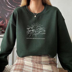 Puede incluir: Sudadera verde oscuro con un gráfico blanco de un sol saliendo sobre las nubes. El texto "even the darkest night will end and the sun will rise" está impreso debajo. La sudadera tiene mangas largas y cuello redondo.