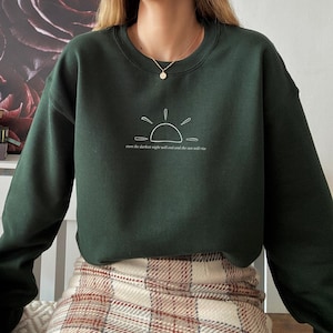 Puede incluir: Sudadera verde oscuro con un gráfico blanco de un sol naciente y el texto "even the darkest night will end and the sun will rise." La sudadera de manga larga tiene cuello redondo.
