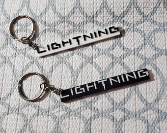 May include: Two keychains with the word "LIGHTNING" printed on them. One keychain is white with black lettering and the other is black with white lettering.