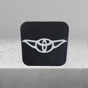 May include: A black square sticker with a white outline of Yoda's ears and the Toyota logo in the centre.