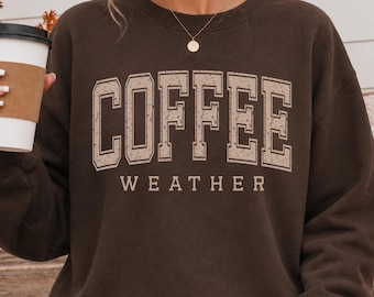 May include: A dark brown crewneck sweatshirt featuring the text COFFEE WEATHER in a distressed, light beige collegiate font. The word COFFEE is displayed in large, bold block letters above the word WEATHER in a smaller, matching font style.
