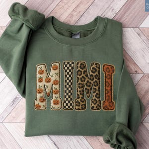 May include: A military green crewneck sweatshirt featuring the word MIMI in large block letters. Each letter has a distinct pattern: the first M has orange pumpkins, the I is black and white checkered, the second M is leopard print, and the I is brown.