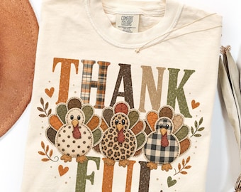 May include: An ivory t-shirt featuring the text THANK FUL in large, distressed block letters. Three turkeys with patterned bodies—polka dot, leopard print, and buffalo plaid—are positioned between the words, accented by small hearts and leaves.