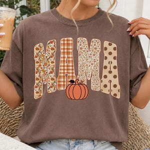 Può includere: Una t-shirt marrone a maniche corte con la parola MAMA stampata in grandi lettere maiuscole. Ogni lettera presenta un motivo diverso, tra cui fiori, quadri arancioni e bianchi e pois marroni. Una zucca arancione è centrata sotto il testo.