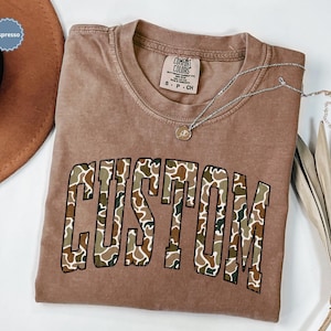 Custom Camo Shirt featuring bold varsity-style font — the perfect Camouflage Shirt for deer hunting, duck hunting, or casual wear during hunting season. This cozy Camo Sweatshirt makes a thoughtful Hunting Lover Gift, combining rugged outdoor style with personalized comfort.