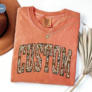Custom Camo Shirt featuring bold varsity-style font — the perfect Camouflage Shirt for deer hunting, duck hunting, or casual wear during hunting season. This cozy Camo Sweatshirt makes a thoughtful Hunting Lover Gift, combining rugged outdoor style with personalized comfort.