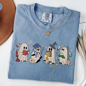 Halloween ghost shirt featuring four cute reading ghosts dressed as magical wizard characters with books, broomstick, owl, potion, and spooky season graphics on a cozy Halloween graphic tee.