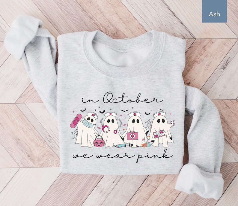 Pink Ghost Nurse Shirt featuring adorable ghost nurses with pink nurse stethoscopes and nurse items, with the phrase "In October We Wear Pink" in a script lettering. This Halloween Tee is perfect for healthcare heroes supporting Breast Cancer Awareness during spooky season. A thoughtful and festive Oncology Nurse Tee or Cute Nurse Tshirt for October events, hospital shifts, or awareness walks. Celebrate with this Pink Ribbon Shirt designed for nurses or medical assistants who care.