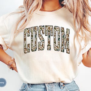 Custom Camo Shirt featuring bold varsity-style font — the perfect Camouflage Shirt for deer hunting, duck hunting, or casual wear during hunting season. This cozy Camo Sweatshirt makes a thoughtful Hunting Lover Gift, combining rugged outdoor style with personalized comfort.
