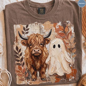 May include: An espresso-colored t-shirt featuring a graphic print of a shaggy brown Highland cow standing next to a white ghost. The design includes textured brushstroke details, autumn-colored leaves, and a cracked background effect. The shirt has a crew neckline and a Comfort Colors brand label at the inner neck.